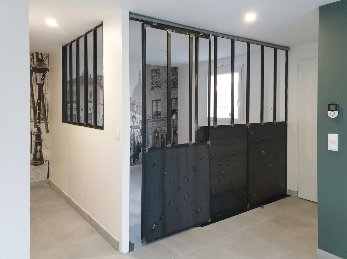 Photographie d'un intérieur résidentiel moderne montrant une cloison vitrée épurée avec cadre en acier ou aluminium gris anthracite, séparant visuellement une cuisine ouverte d'un salon living, laissant passer la lumière naturelle tout en créant une délimitation architecturale claire. La verrière doit être le sujet principal de l'image, mettant en évidence sa structure métallique et son intégration harmonieuse dans l'espace, avec les deux zones (cuisine et salon) visibles et bien éclairées par la lumière du jour.